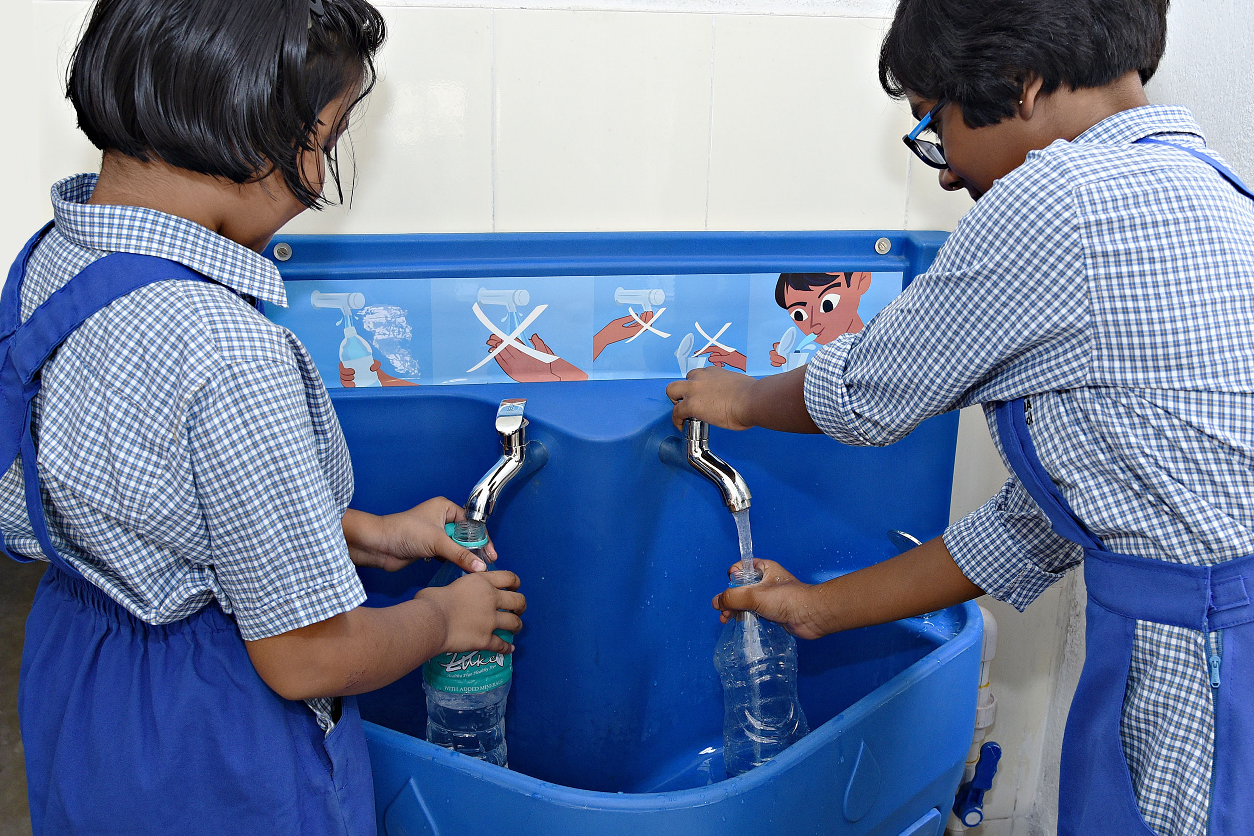 Two young girls in India wearing school uniforms and filling plastic water bottles from a blue Splash drinking water station.